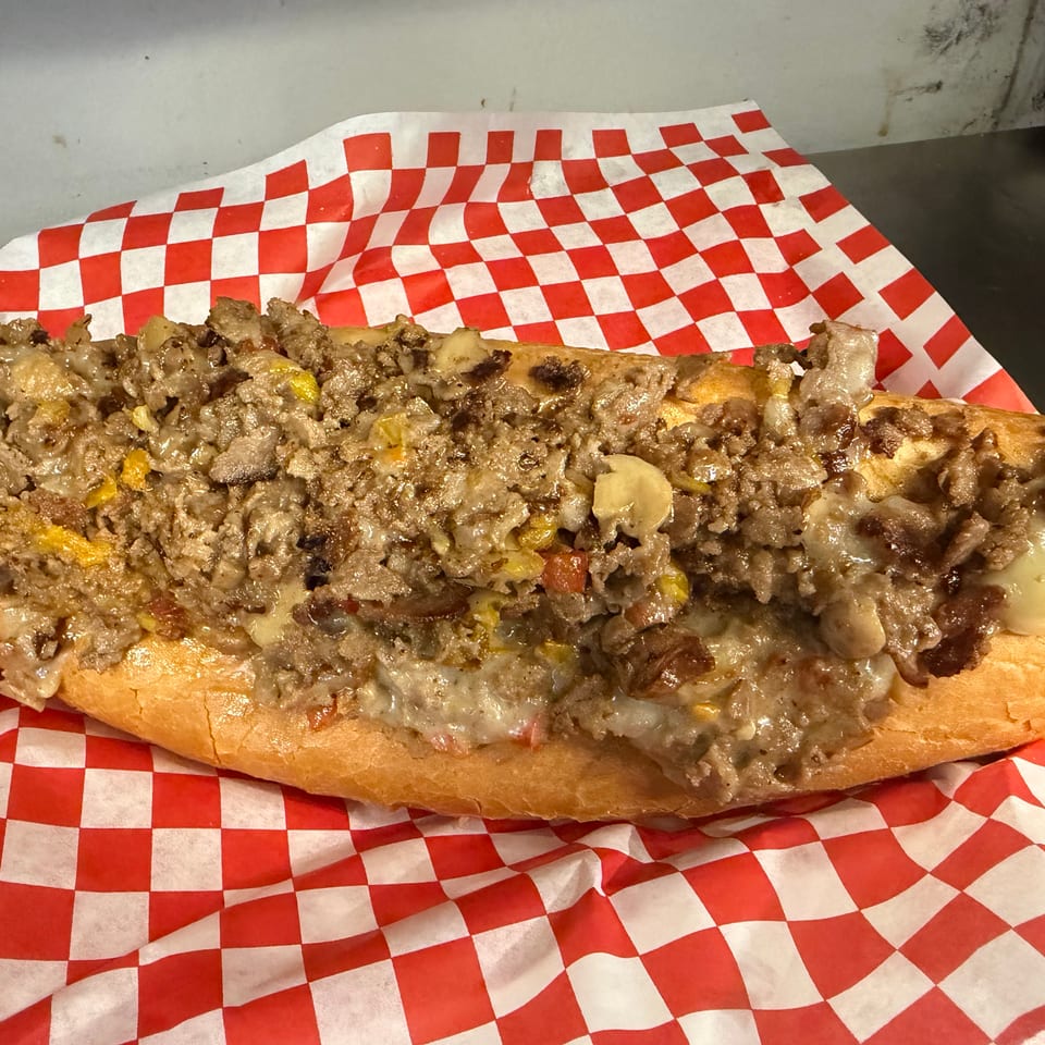 Loaded cheesesteak sandwich with chopped beef, melted cheese, peppers, and onions on a hoagie roll atop red-and-white checkered paper.