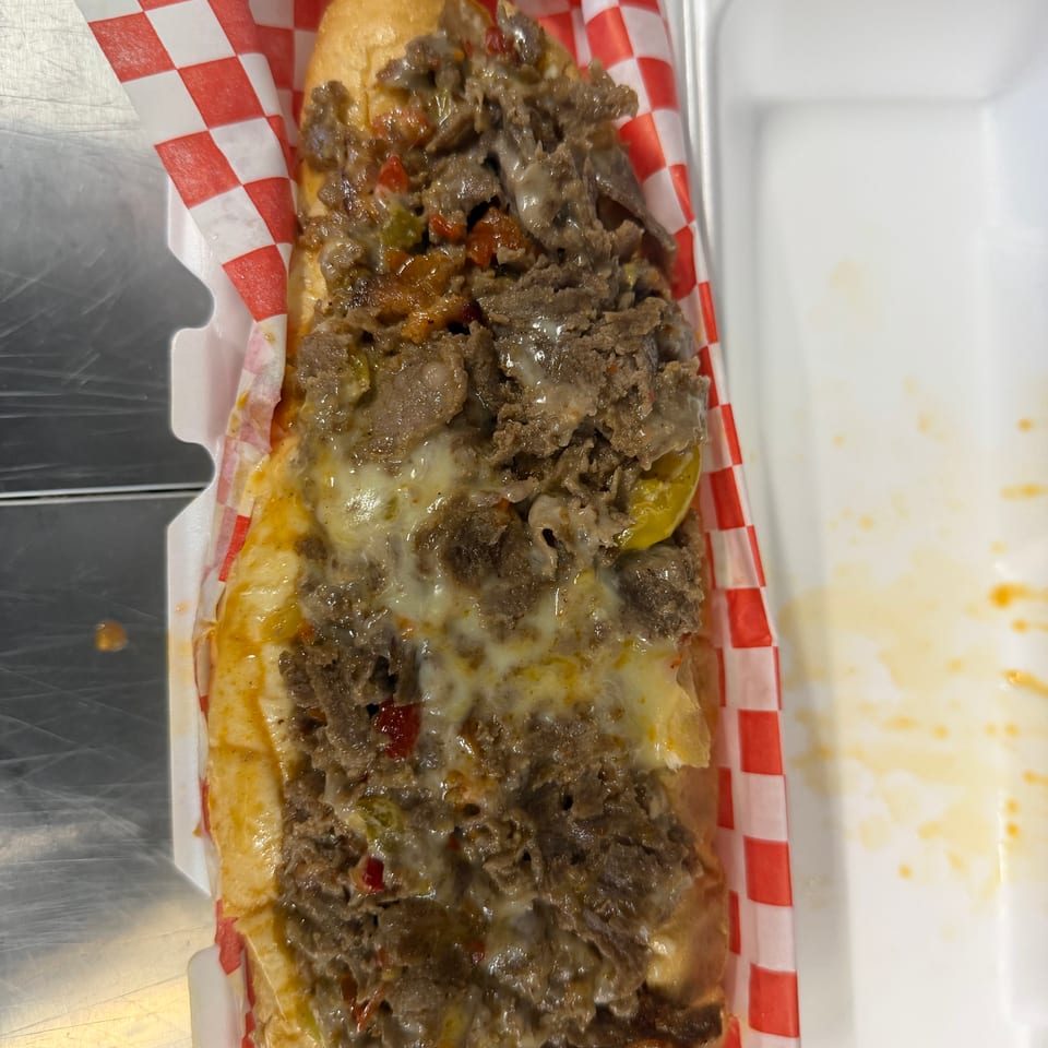 Loaded cheesesteak sandwich with sliced beef, melted cheese, peppers, onions, and tomatoes in a checkered paper-lined tray.
