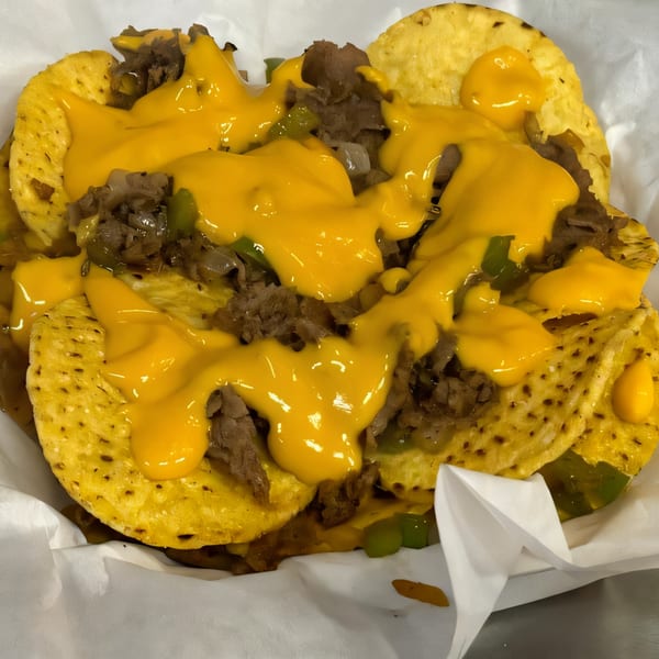 Cheesesteak nachos with melted cheese, steak, onions, and green peppers in a white paper container.