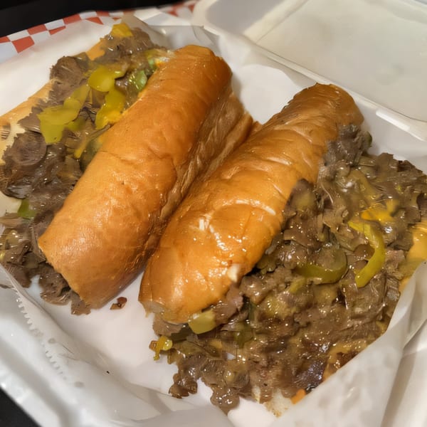 Two Philly Boyz cheesesteaks with melted cheese and peppers in a white takeout container.